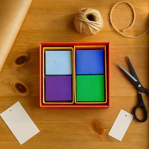 May include: A top-down view of a gift box with four colorful square compartments. The compartments contain blue, purple, and green items. A ball of twine, scissors, and blank tags are on a wooden surface.