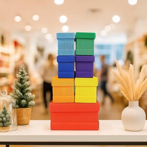May include: A stack of colorful gift boxes in shades of red, orange, yellow, blue, green, and purple. The boxes are arranged on a white surface, with a blurred background of a store interior. A small Christmas tree and vase are also visible.