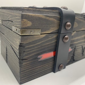 K&ouml;nnte beinhalten: Eine Holzkiste mit einem schwarzen Lederriemen und Metallverzierungen. Die Kiste hat einen roten Streifen, der an der Seite bemalt ist, und einen Metallverschluss an der Vorderseite.