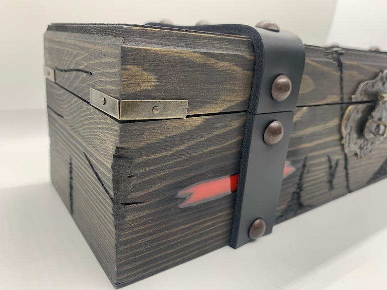 K&ouml;nnte beinhalten: Eine Holzkiste mit einem schwarzen Lederriemen und Metallverzierungen. Die Kiste hat einen roten Streifen, der an der Seite bemalt ist, und einen Metallverschluss an der Vorderseite.