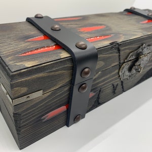 K&ouml;nnte beinhalten: Eine Holzkiste mit einem schwarzen Lederriemen und Metallverzierungen. Die Kiste hat ein rot-schwarzes Design und einen Metallverschluss.