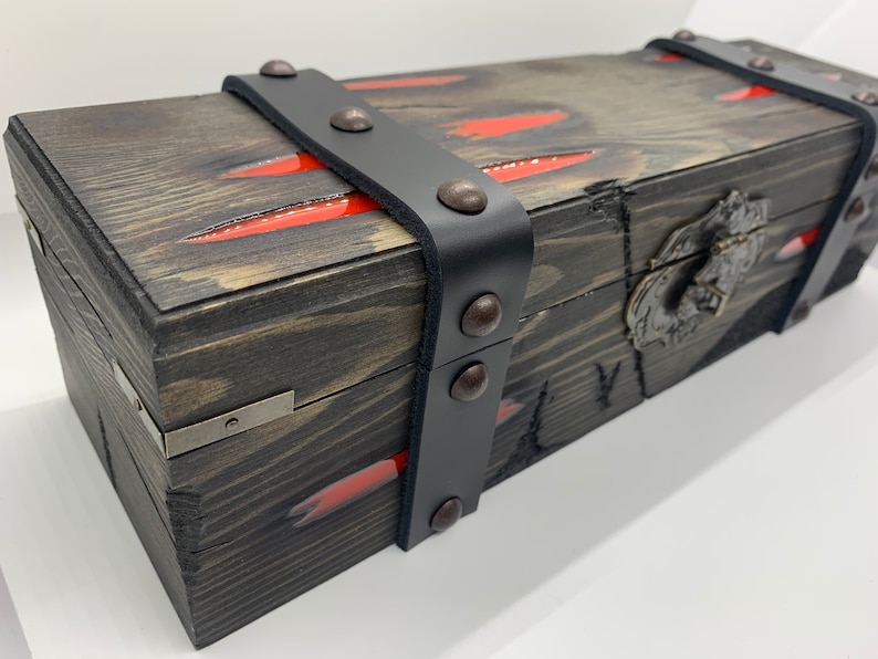 K&ouml;nnte beinhalten: Eine Holzkiste mit einem schwarzen Lederriemen und Metallverzierungen. Die Kiste hat ein rot-schwarzes Design und einen Metallverschluss.