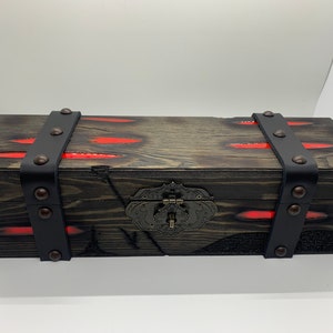 K&ouml;nnte beinhalten: Eine Holzkiste mit einem schwarzen Lederriemen und Metallverzierungen. Die Kiste hat eine rote Harzeinlage und einen Metallverschluss.