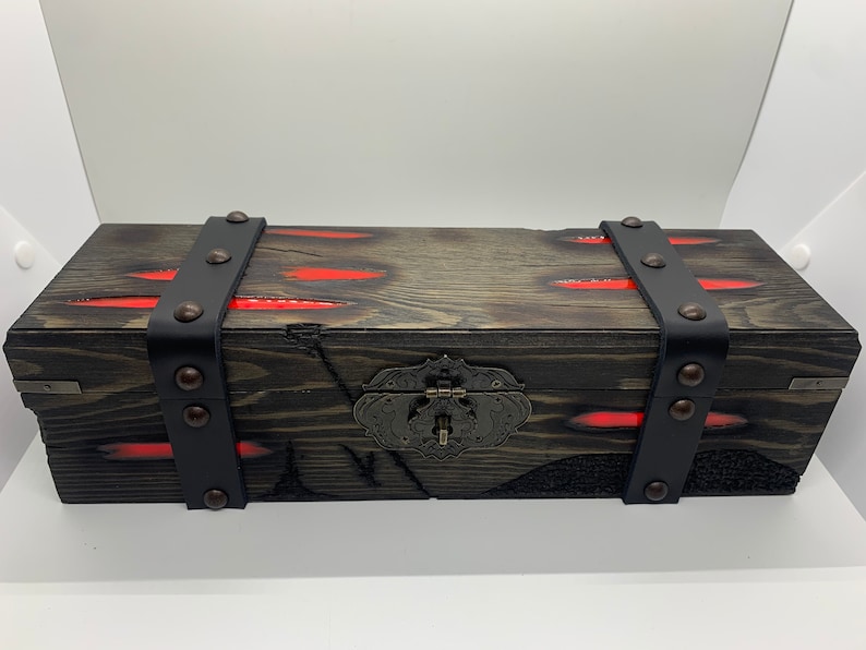 K&ouml;nnte beinhalten: Eine Holzkiste mit einem schwarzen Lederriemen und Metallverzierungen. Die Kiste hat eine rote Harzeinlage und einen Metallverschluss.