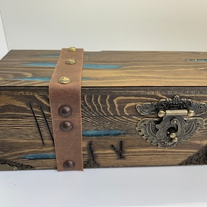 May include: A wooden box with a brown leather strap and metal accents. The box has a decorative metal latch and is stained a dark brown color.