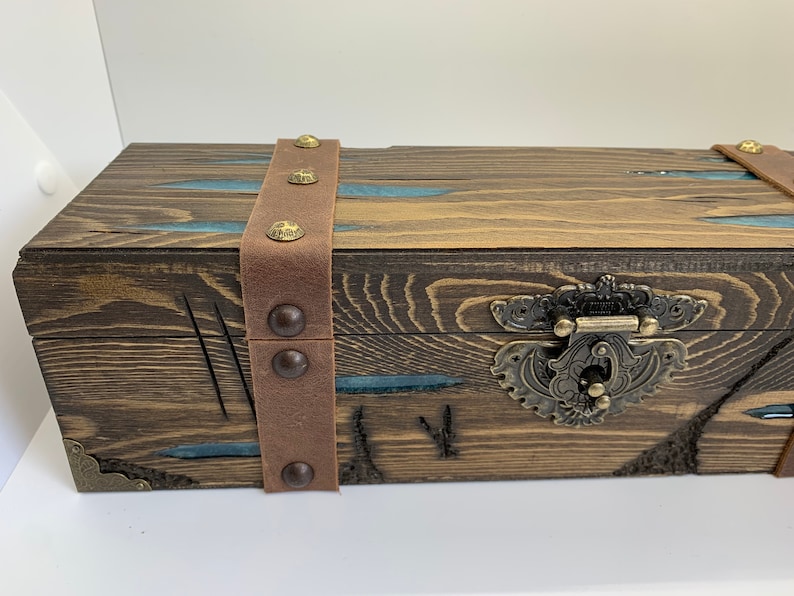 May include: A wooden box with a brown leather strap and metal accents. The box has a decorative metal latch and is stained a dark brown color.
