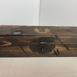 K&ouml;nnte beinhalten: Eine Holzkiste mit einer dunkelbraunen Beize und einem Messingverschluss. Die Oberseite der Kiste hat ein goldenes Emblem mit einem stilisierten Vogelmotiv.