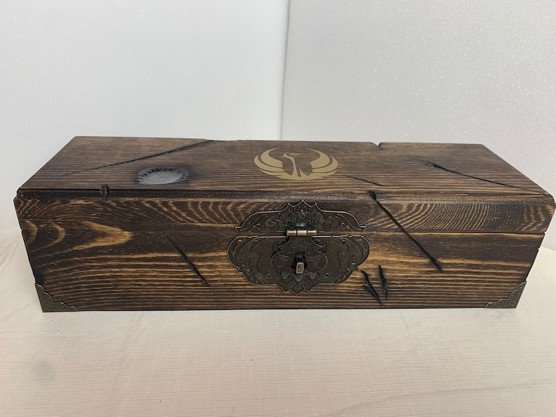 K&ouml;nnte beinhalten: Eine Holzkiste mit einer dunkelbraunen Beize und einem Messingverschluss. Die Oberseite der Kiste hat ein goldenes Emblem mit einem stilisierten Vogelmotiv.
