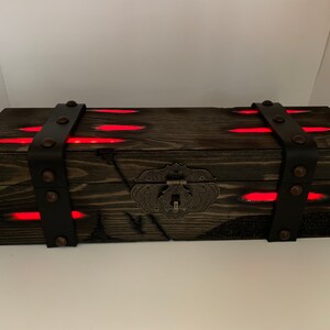 K&ouml;nnte beinhalten: Eine dunkle Holzkiste mit einem Metallverschluss und Lederriemen. Die Kiste wird von innen mit roten Lichtern beleuchtet.