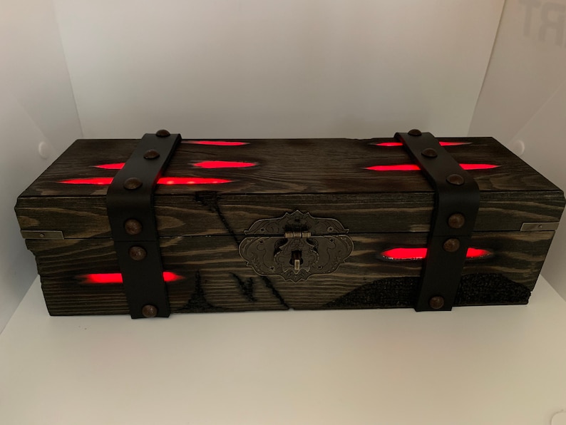K&ouml;nnte beinhalten: Eine dunkle Holzkiste mit einem Metallverschluss und Lederriemen. Die Kiste wird von innen mit roten Lichtern beleuchtet.