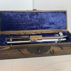 K&ouml;nnte beinhalten: Ein silbernes Lichtschwert mit goldener Akzent ist in einer Holzkiste mit blauer Samtf&uuml;tterung ausgestellt. Die Kiste hat einen dekorativen Metallverschluss.