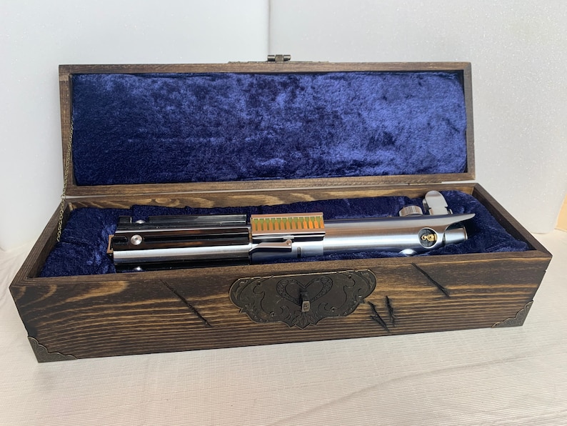 K&ouml;nnte beinhalten: Ein silbernes Lichtschwert mit goldener Akzent ist in einer Holzkiste mit blauer Samtf&uuml;tterung ausgestellt. Die Kiste hat einen dekorativen Metallverschluss.