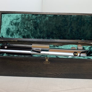 Pode incluir: Um sabre de luz prateado com cabo preto e detalhes dourados &eacute; exibido em uma caixa de madeira com forro de veludo verde.