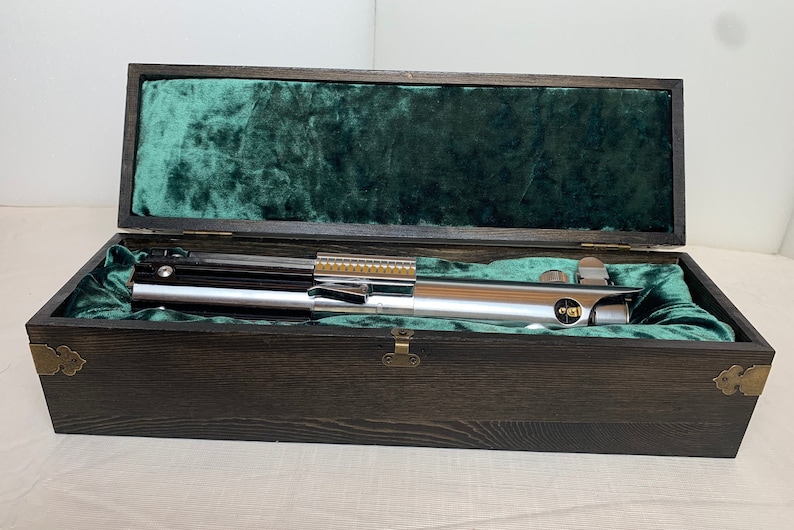 Pode incluir: Um sabre de luz prateado com cabo preto e detalhes dourados &eacute; exibido em uma caixa de madeira com forro de veludo verde.