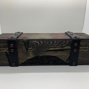 K&ouml;nnte beinhalten: Eine Holzkiste mit schwarzen Lederriemen und Metallverzierungen. Die Kiste hat eine dunkle Holzoptik und einen Used-Look. Die Riemen sind mit Metallnieten befestigt.