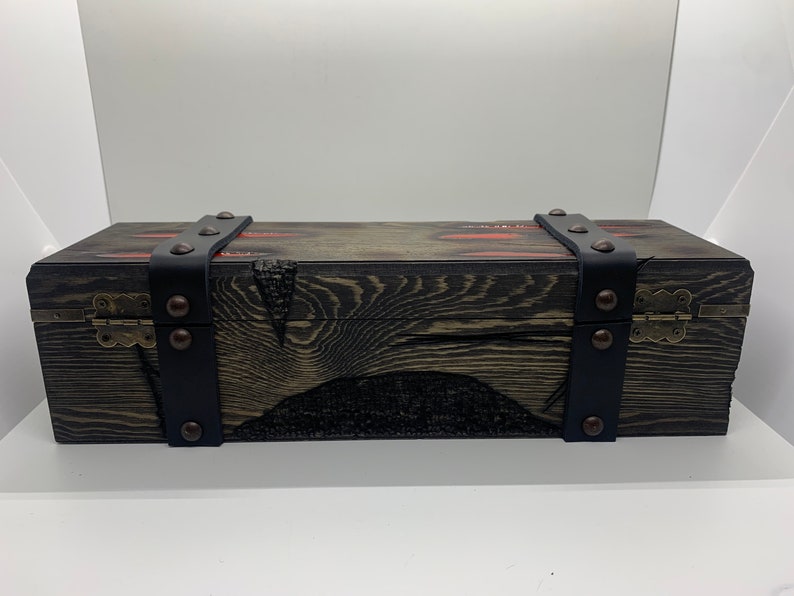 K&ouml;nnte beinhalten: Eine Holzkiste mit schwarzen Lederriemen und Metallverzierungen. Die Kiste hat eine dunkle Holzoptik und einen Used-Look. Die Riemen sind mit Metallnieten befestigt.