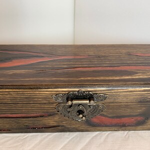 May include: A wooden box with a dark brown stain and red accents. The box has a metal latch with an ornate design.