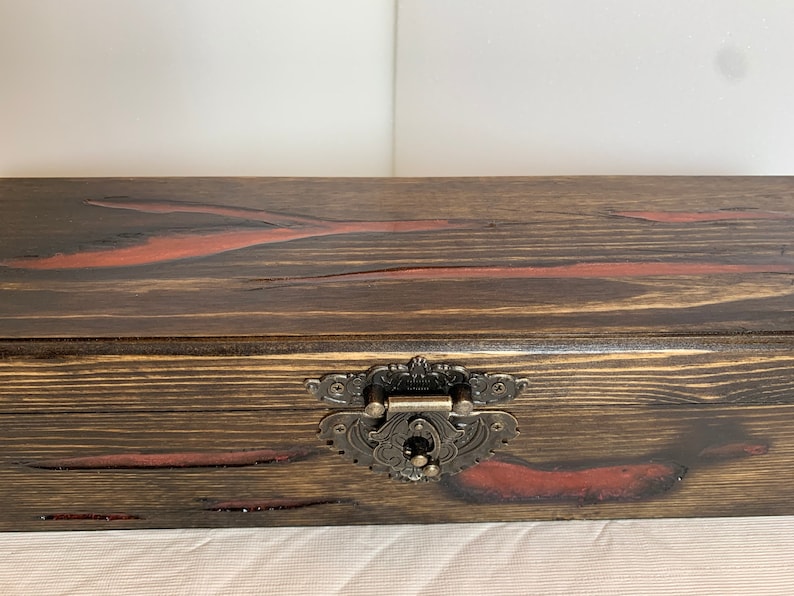 May include: A wooden box with a dark brown stain and red accents. The box has a metal latch with an ornate design.