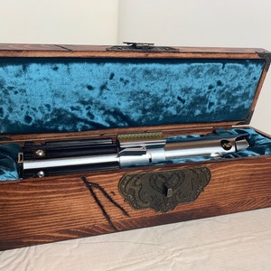 May include: A silver lightsaber with a black handle is displayed in a wooden box with a blue velvet lining. The box has a brass latch and decorative metal accents.