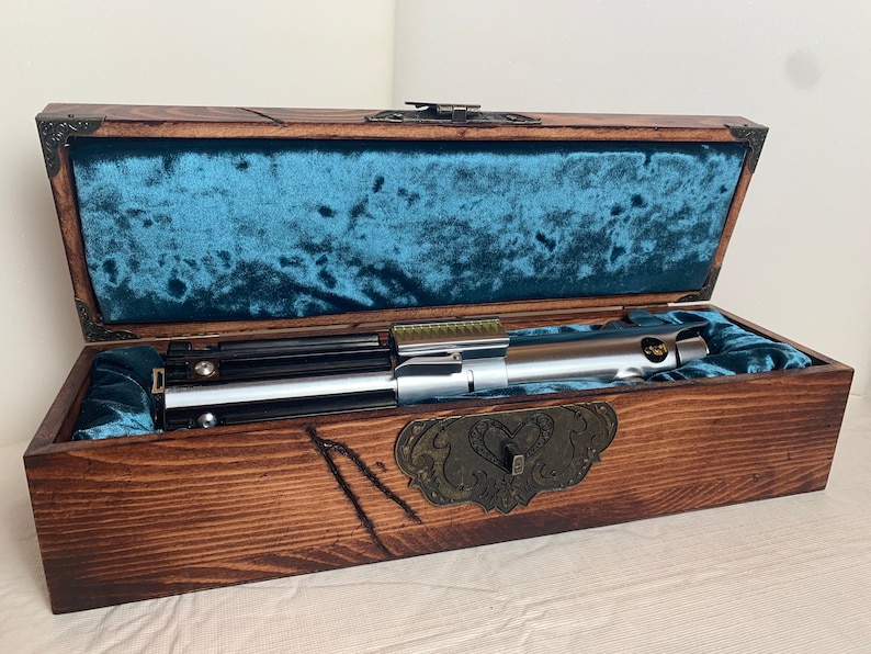 May include: A silver lightsaber with a black handle is displayed in a wooden box with a blue velvet lining. The box has a brass latch and decorative metal accents.