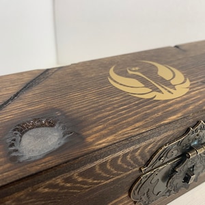 K&ouml;nnte beinhalten: Eine Holzkiste mit einem goldenen Rebellenallianz-Symbol auf dem Deckel. Die Kiste hat einen Metallverschluss und eine abgenutzte Oberfl&auml;che.