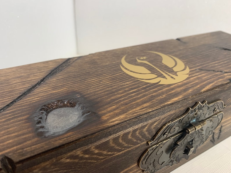 K&ouml;nnte beinhalten: Eine Holzkiste mit einem goldenen Rebellenallianz-Symbol auf dem Deckel. Die Kiste hat einen Metallverschluss und eine abgenutzte Oberfl&auml;che.