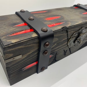 K&ouml;nnte beinhalten: Eine Holzkiste mit einem schwarzen Lederriemen und Metallverzierungen. Die Kiste hat rote Streifen auf der Oberseite und einen Metallverschluss.