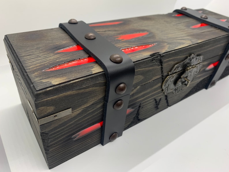 K&ouml;nnte beinhalten: Eine Holzkiste mit einem schwarzen Lederriemen und Metallverzierungen. Die Kiste hat rote Streifen auf der Oberseite und einen Metallverschluss.