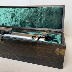 Pode incluir: Um sabre de luz prateado em uma caixa de madeira com um forro de veludo verde. A caixa tem dobradi&ccedil;as de lat&atilde;o e uma trava de lat&atilde;o.