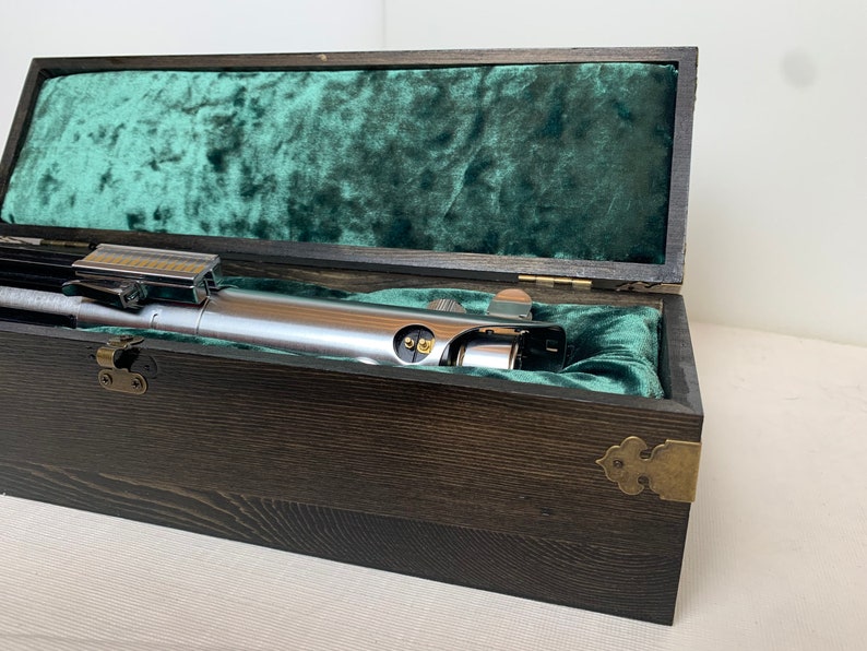 Pode incluir: Um sabre de luz prateado em uma caixa de madeira com um forro de veludo verde. A caixa tem dobradi&ccedil;as de lat&atilde;o e uma trava de lat&atilde;o.