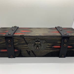 K&ouml;nnte beinhalten: Eine Holzkiste mit einem schwarzen Lederriemen und Metallverzierungen. Die Kiste hat eine abgenutzte Oberfl&auml;che und rote Akzente.