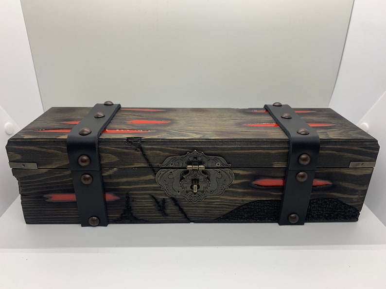 K&ouml;nnte beinhalten: Eine Holzkiste mit einem schwarzen Lederriemen und Metallverzierungen. Die Kiste hat eine abgenutzte Oberfl&auml;che und rote Akzente.