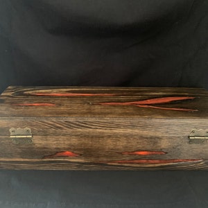 May include: A wooden box with a dark brown stain and red resin accents. The box has two brass hinges on the front.