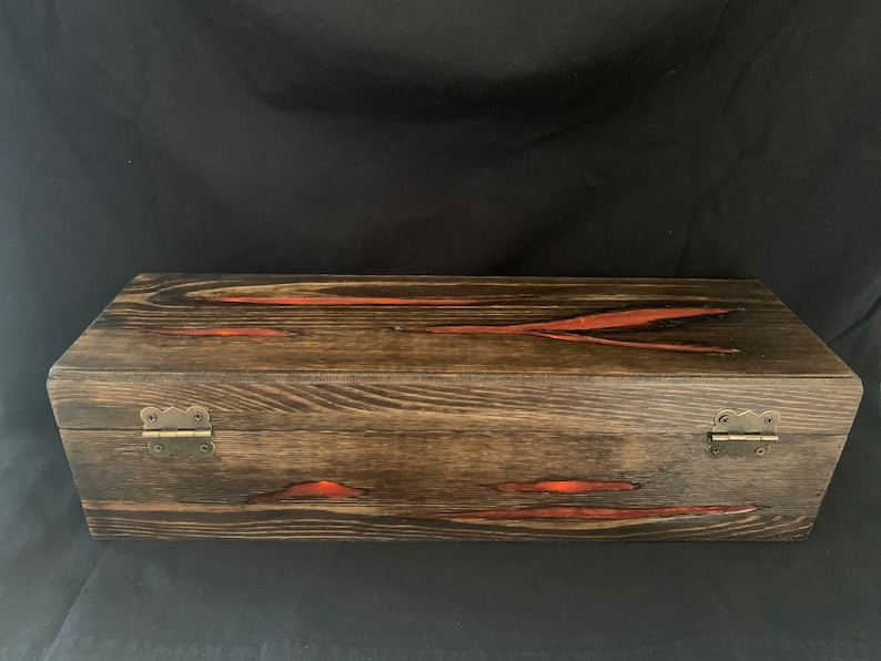 May include: A wooden box with a dark brown stain and red resin accents. The box has two brass hinges on the front.