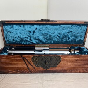 May include: A wooden box with a blue velvet lining, containing a silver lightsaber. The box has a brass latch and decorative carvings.