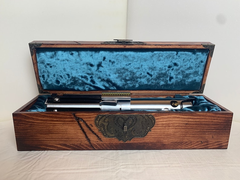 May include: A wooden box with a blue velvet lining, containing a silver lightsaber. The box has a brass latch and decorative carvings.