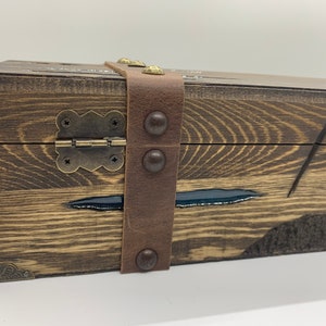 May include: A wooden box with a leather strap and metal accents. The box has a dark wood finish and a black resin inlay. The box is closed and the strap is fastened.