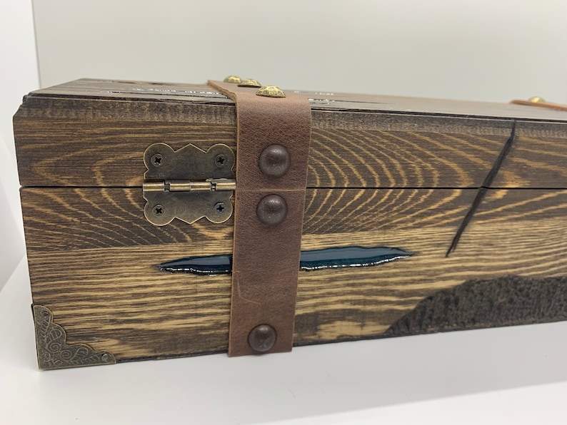 May include: A wooden box with a leather strap and metal accents. The box has a dark wood finish and a black resin inlay. The box is closed and the strap is fastened.