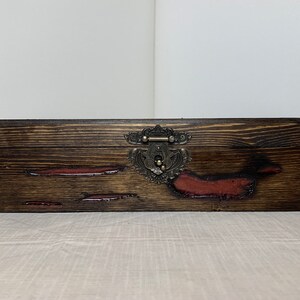 May include: A wooden box with a dark brown stain and red accents. The box has a metal latch with an ornate design.