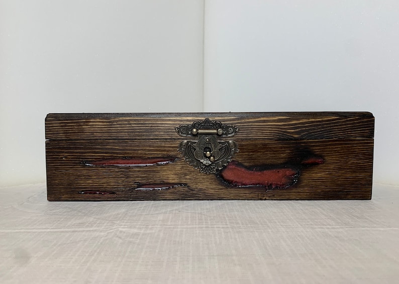 May include: A wooden box with a dark brown stain and red accents. The box has a metal latch with an ornate design.