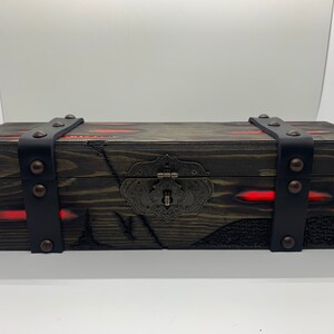 K&ouml;nnte beinhalten: Eine Holzkiste mit einem schwarzen Lederriemen und Metallverzierungen. Die Kiste hat eine dunkelbraune Holzoptik mit roten Akzenten und einem Metallverschluss.