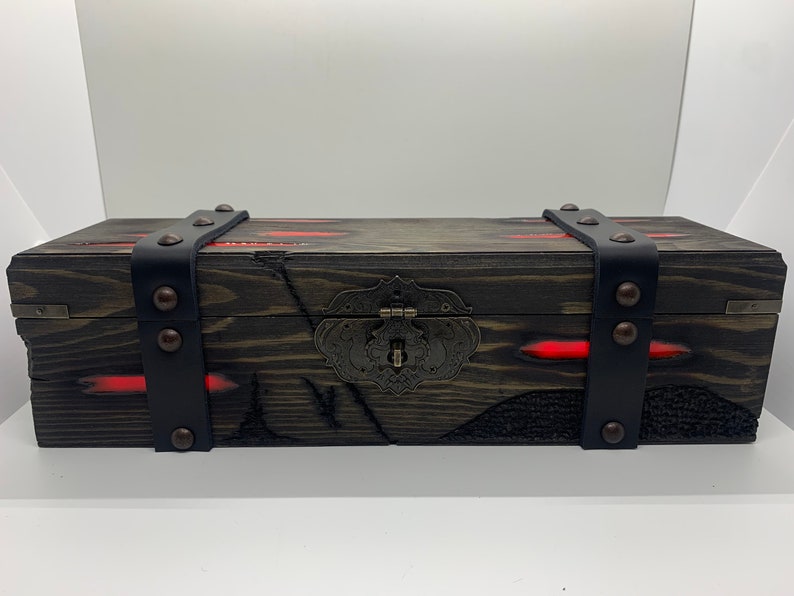 K&ouml;nnte beinhalten: Eine Holzkiste mit einem schwarzen Lederriemen und Metallverzierungen. Die Kiste hat eine dunkelbraune Holzoptik mit roten Akzenten und einem Metallverschluss.
