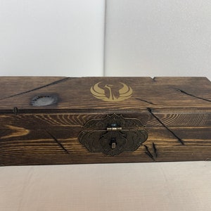 K&ouml;nnte beinhalten: Eine Holzkiste mit einer dunkelbraunen Beize und einem goldenen Emblem auf dem Deckel. Die Kiste hat einen Metallverschluss und Scharniere.