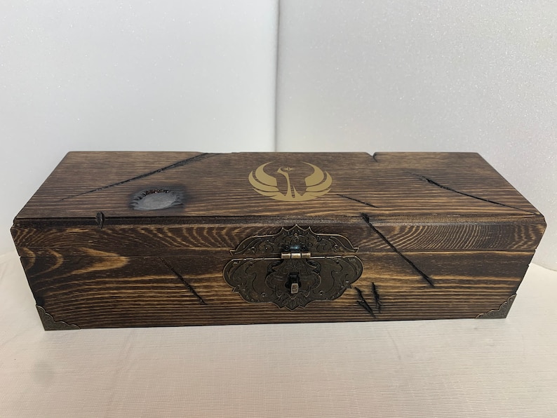 K&ouml;nnte beinhalten: Eine Holzkiste mit einer dunkelbraunen Beize und einem goldenen Emblem auf dem Deckel. Die Kiste hat einen Metallverschluss und Scharniere.