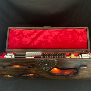 May include: A wooden box with a red velvet interior and a silver and black lightsaber inside. The box has a decorative metal latch and a glowing orange light emanating from the bottom.