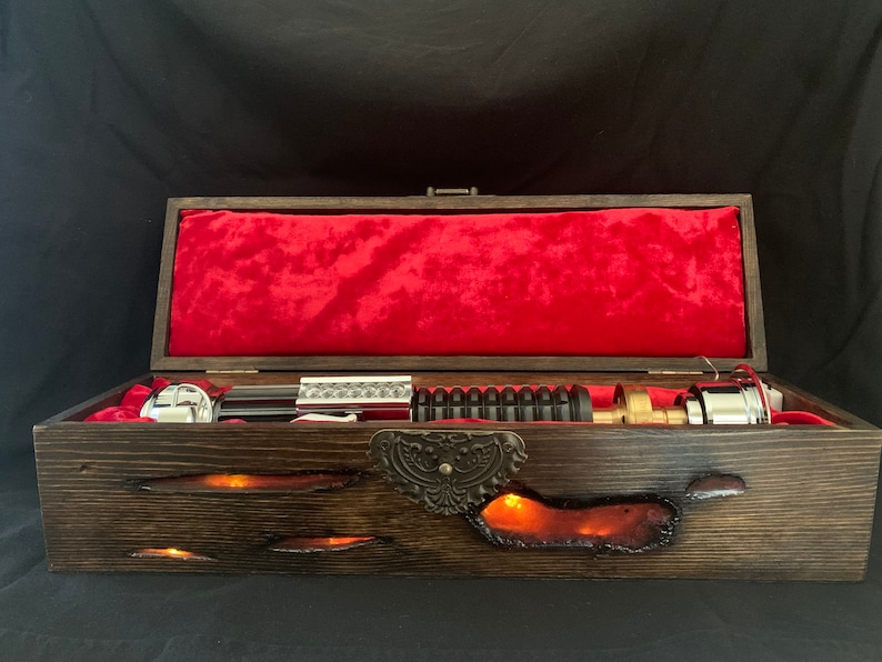 May include: A wooden box with a red velvet interior and a silver and black lightsaber inside. The box has a decorative metal latch and a glowing orange light emanating from the bottom.