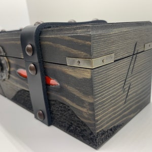 K&ouml;nnte beinhalten: Eine Holzkiste mit einem schwarzen Lederriemen und Metallverzierungen. Die Kiste hat eine dunkelbraune Holzoptik und einen roten Akzent.