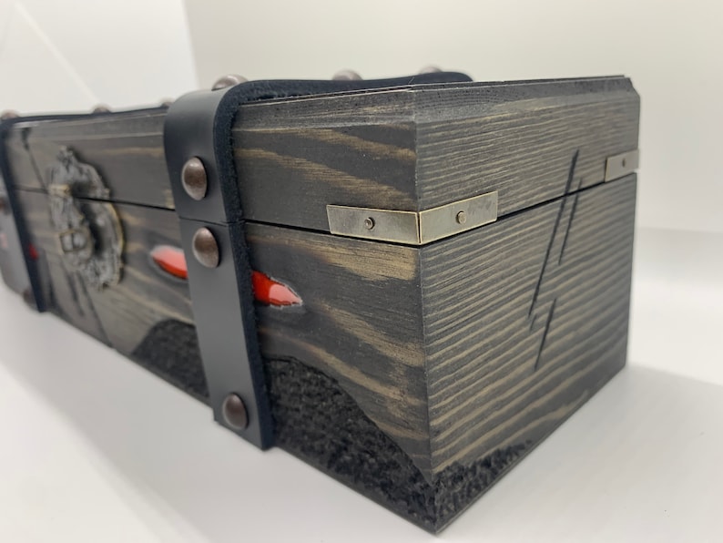 K&ouml;nnte beinhalten: Eine Holzkiste mit einem schwarzen Lederriemen und Metallverzierungen. Die Kiste hat eine dunkelbraune Holzoptik und einen roten Akzent.