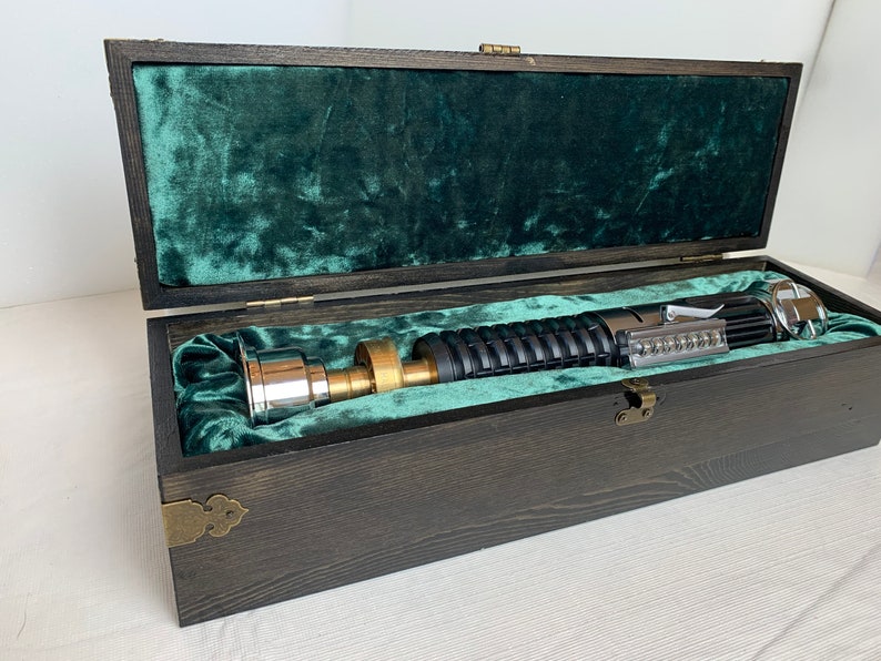 Pode incluir: Um sabre de luz numa caixa de madeira com um forro de veludo verde. O sabre de luz &eacute; prateado e preto com uma pega de lat&atilde;o.