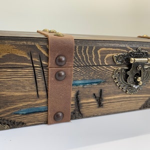 May include: A wooden box with a brown leather strap and metal accents. The box has a carved design and a blue resin inlay.